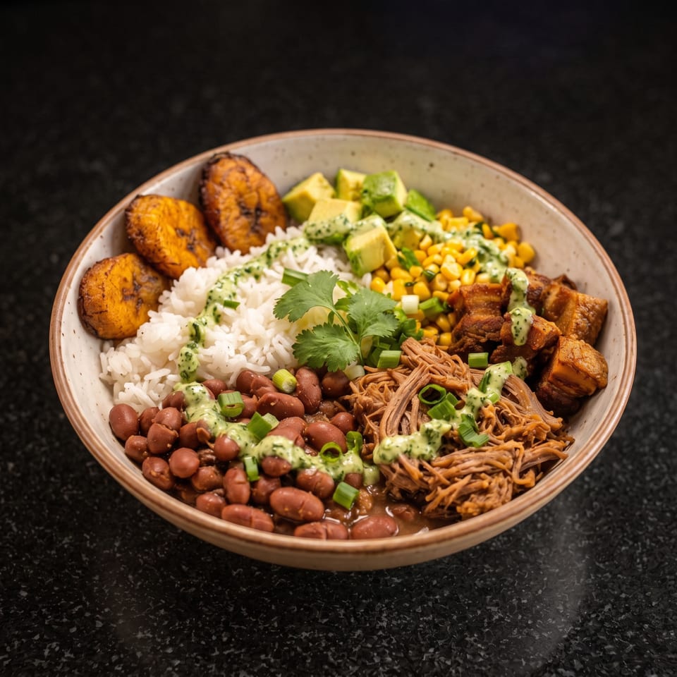 Bowl of Latin-inspired food with rice, beans, plantains, avocado, corn, shredded meat, crispy pork, and cilantro sauce.