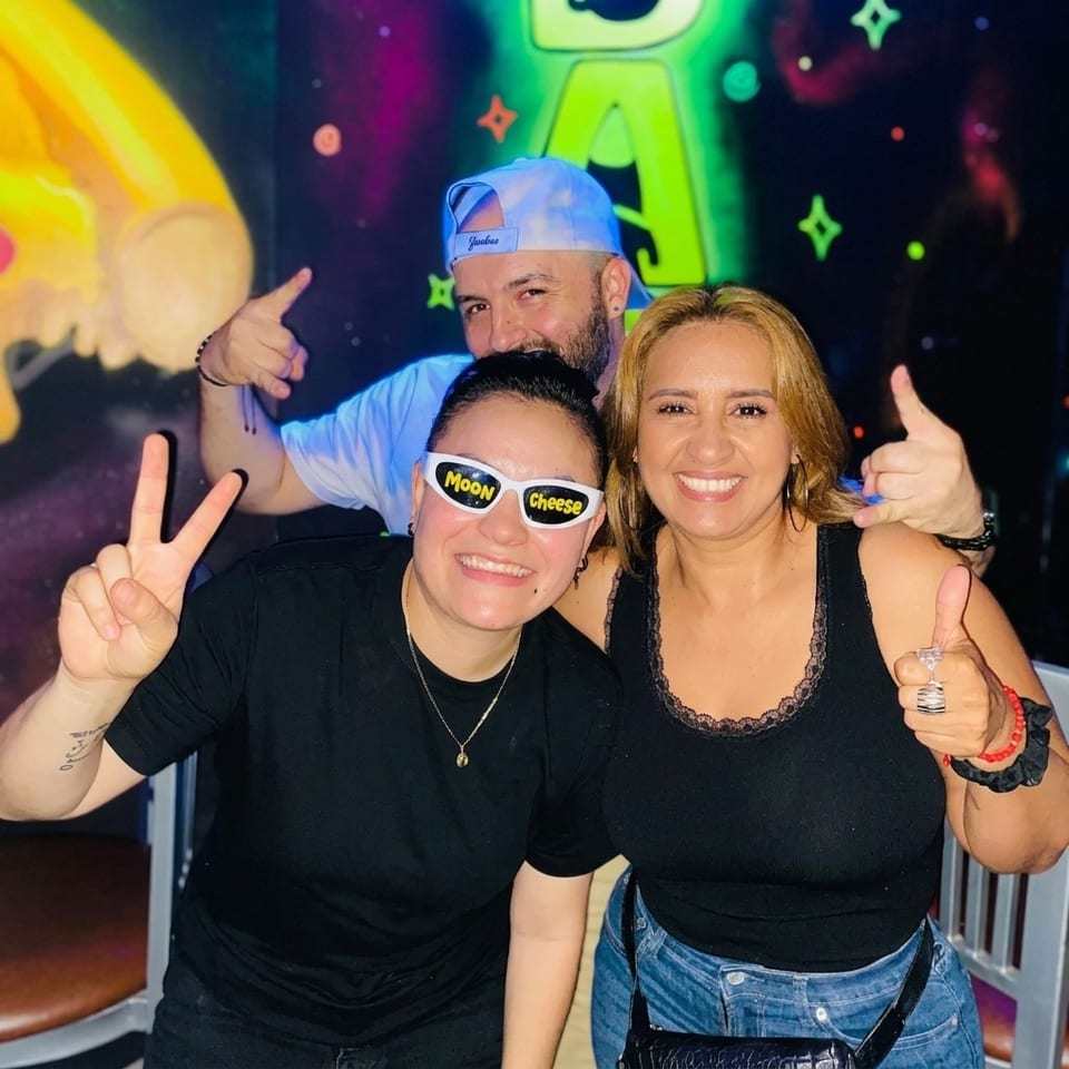 Three smiling people pose together in a colorful neon-lit venue, giving thumbs-up and peace signs.