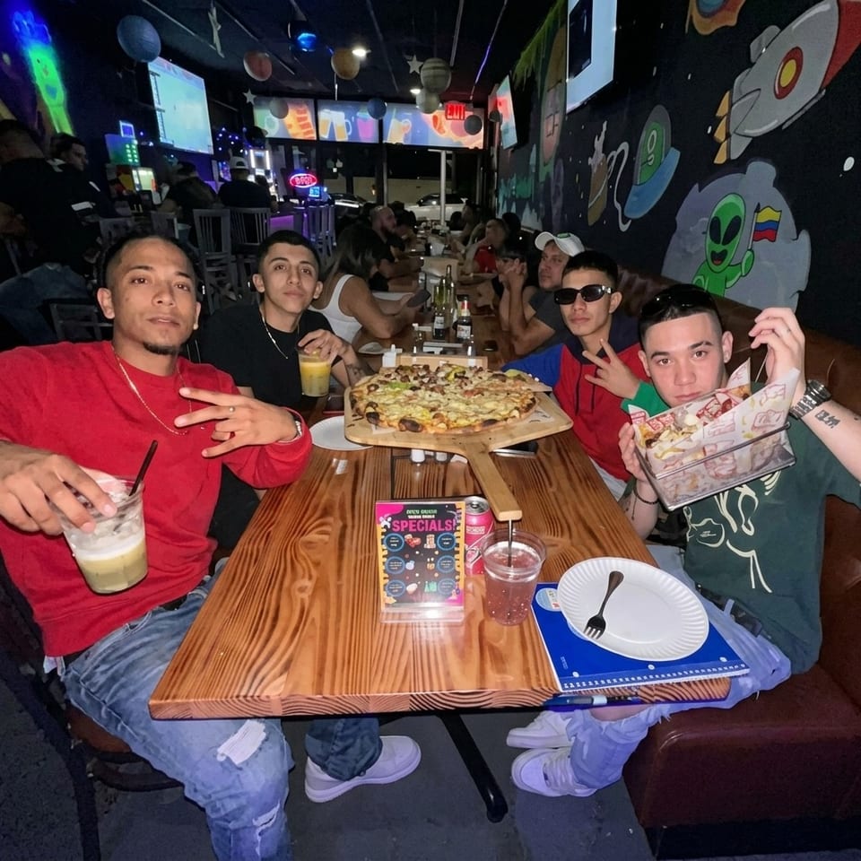 Friends pose around a table with pizza and drinks inside a lively, space-themed restaurant decorated with colorful murals.