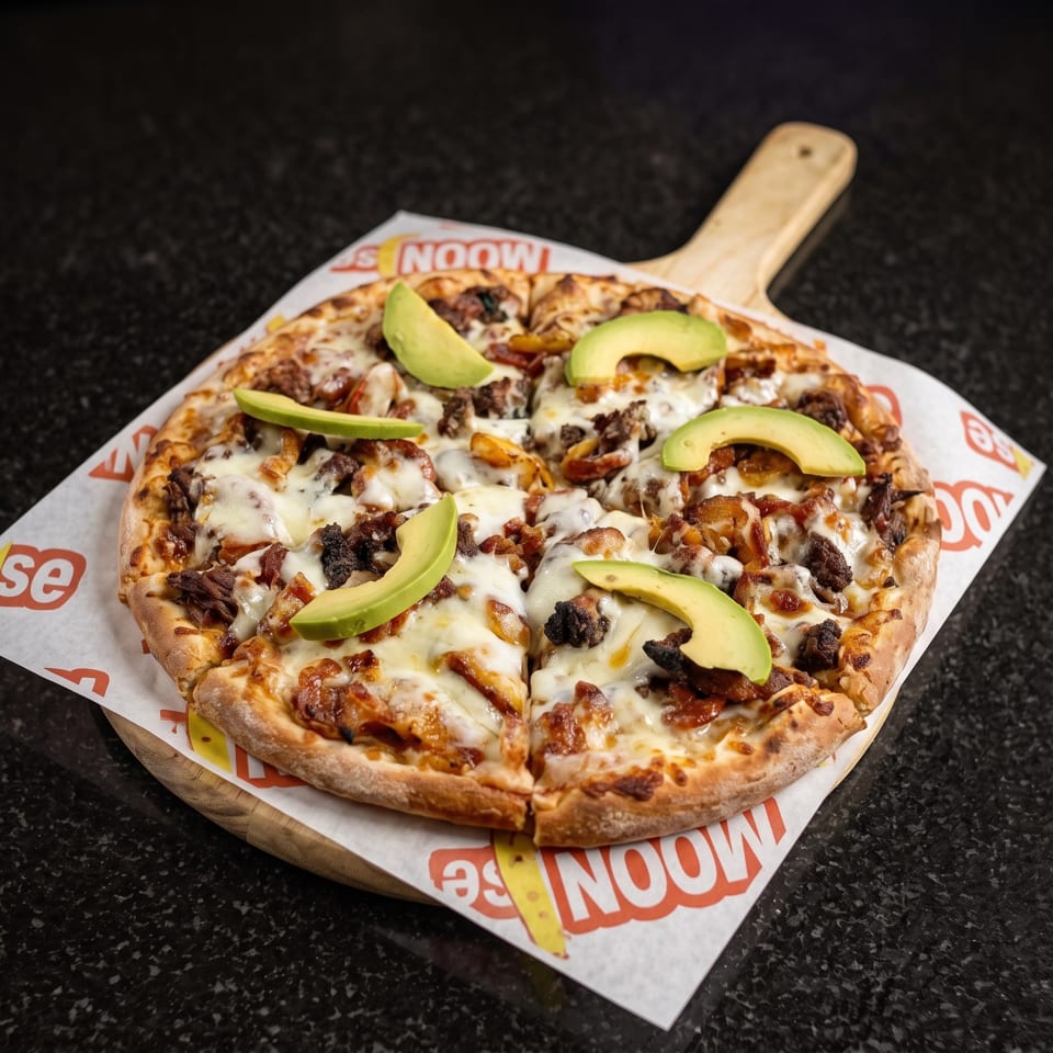 Cheesy meat-topped pizza garnished with avocado slices, served on branded paper atop a wooden pizza board.