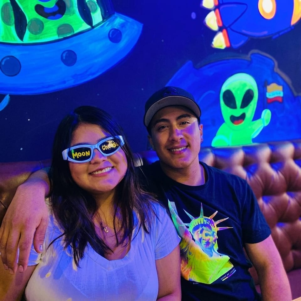 Smiling couple seated in a neon-lit restaurant booth, with colorful alien-themed murals and playful sunglasses.