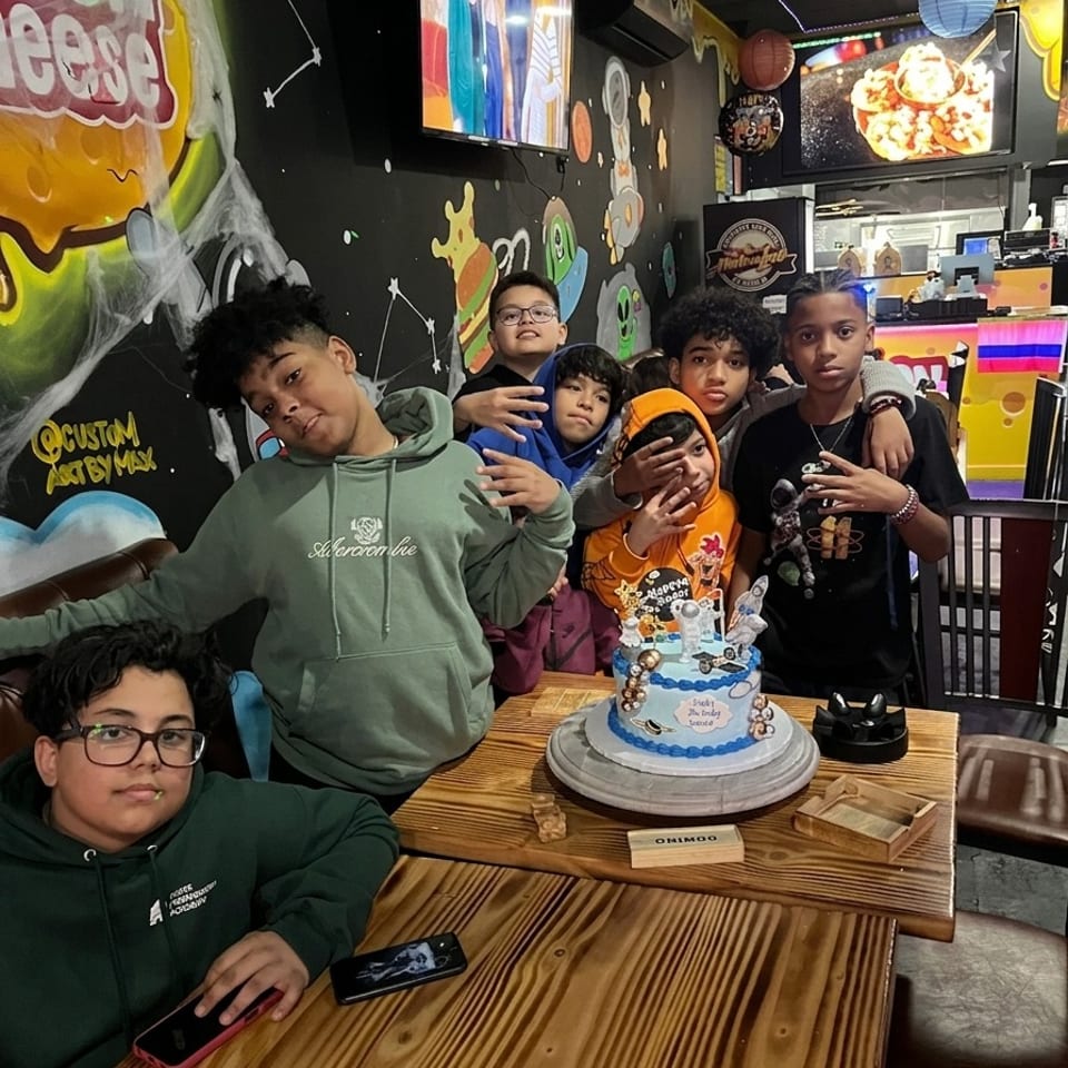 Six children gather around a decorated birthday cake at a colorful, space-themed restaurant table.