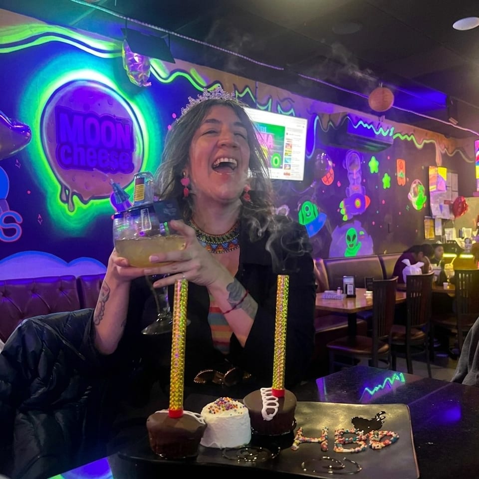 Smiling woman holds a cocktail beside decorated birthday desserts in a colorful, neon-lit Moon Cheese restaurant.