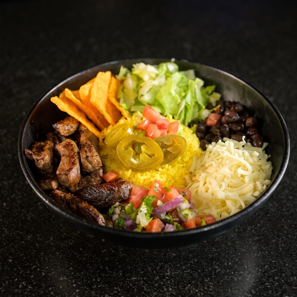 Bowl of grilled steak, yellow rice, black beans, lettuce, tortilla chips, cheese, jalapeños, tomatoes, and salsa.