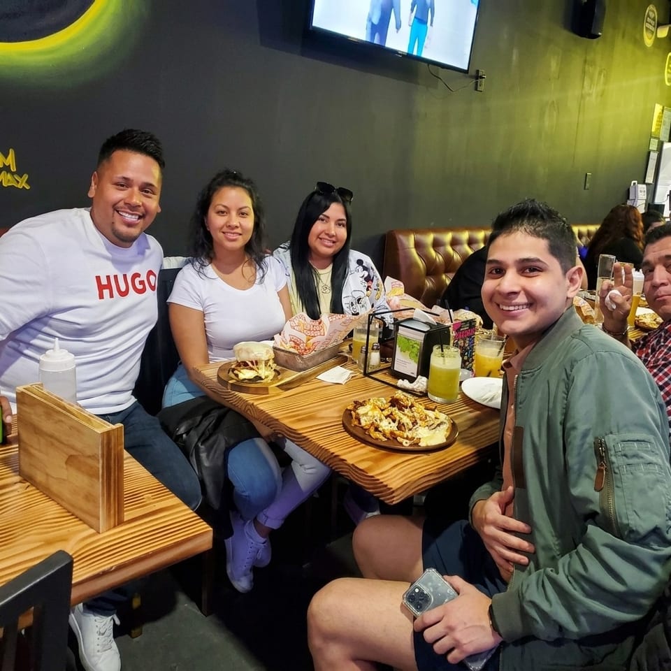 Five friends smile around a restaurant table filled with plated dishes and colorful drinks in a dimly lit dining room.